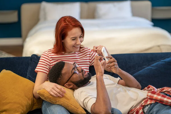 Young diverse couple boyfriend and girlfriend use smartphone while resting on weekend at home, diverse family man and woman look at mobile phone screen choose hotel for vacation