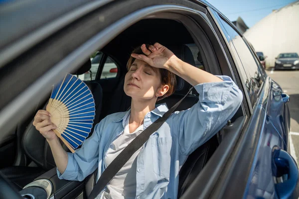 stock image Exhausted tired middle aged woman drives car waves blue fan suffers from stuffiness stands in urban traffic jam in summer hot weather. Overheating, high temperature in car with broken air conditioner.