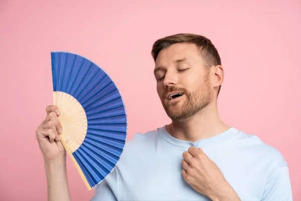 stock image Pleased guy wave reduce heat with hand fan enjoy cooling. Happy satisfied middle aged man with close eyes in light blue t-shirt use waver feel hot no air condition isolated on pink studio background