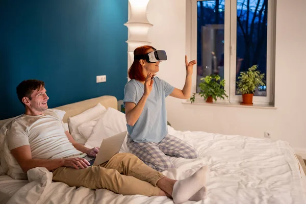 Man watching woman in virtual reality headset. Female enjoys virtual reality with VR headset, smiling admiringly and amazed in company of husband sharing pleasure of modern technologies.
