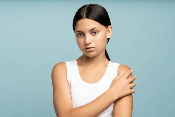 Shy modest teenage girl on studio blue background looking at camera hugging herself one hand. Calm thoughtful pleasant brunette teen girl with ponytail. Woman beauty, youth, adolescence