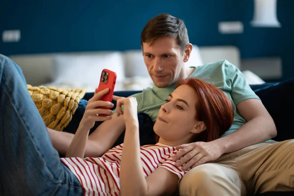 Contented well-rested couple relaxing lying on sofa home resting using smartphone online shopping and video chatting. Married middle aged man and woman choose services on internet book a holiday hotel