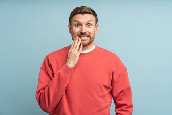 Funny shocked man covering mouth with hand laughs making big eyes, oops, absurd accident. Frightened guy in red sweater opened mouth in surprise, noticing random mistake isolated on blue background.