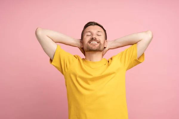 Well-rested smiling man on pink studio background. Carefree happy guy with hands behind neck in open relaxed pose smiles broadly, closed eyes. Contentment, happiness, satisfaction, euphoria, ecstasy.