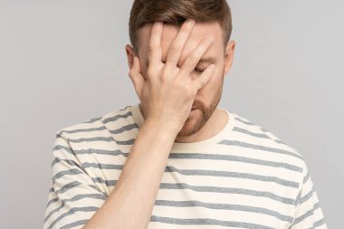Frustrated man lowering head and eyes covers face with hand. Disappointed stressed middle aged male making facepalm gesture with hand, blaming himself for mistake isolated on studio gray background clipart