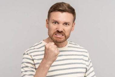 Angry aggressive bearded man clenched fist has evil face looking at camera on grey background. Madly furious guy viciously threatens assault, reprisal or attack. Sincere negative emotions concept. clipart