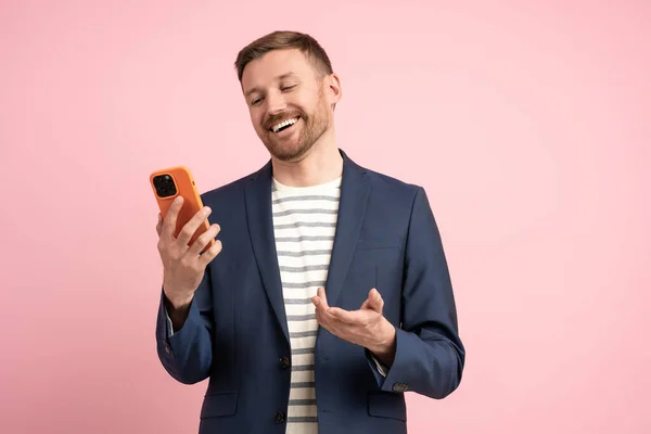 Joyful smiling man looking at mobile phone screen laughing rejoicing at good offer approval of deal over isolated pink background. Delighted trader with smartphone business communicates via video link