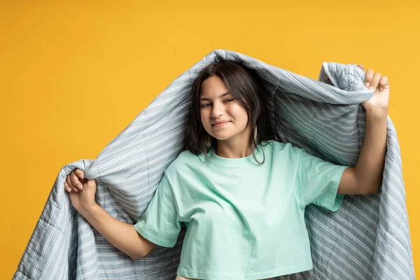 stock image Brunette positive teen girl covering in grey striped blanket on yellow background looking at camera. Advertisement banner poster with smiling girl feeling comfort. Pleasure, resting, relaxing concept.