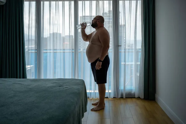 Thirsty overweight man feeling dehydration drinking water in hotel room in hot humid weather standing near window. Fat guy with naked torso suffers from heat, heat stroke, overheating, excess weight.