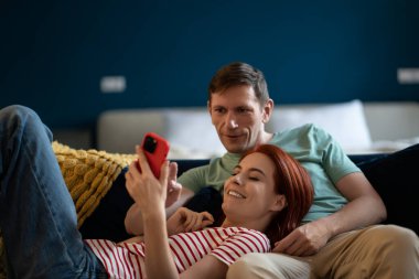 Aşık bir çift rahat koltukta dinleniyor akıllı telefon uygulamalarını kullanarak eğleniyor, uzak video görüşmesinin tadını çıkarıyor, karı kocaya ilginç bir web sitesi gösteriyor, iyi hizmetleri seçiyor, evli çiftler online alışveriş yapıyor.