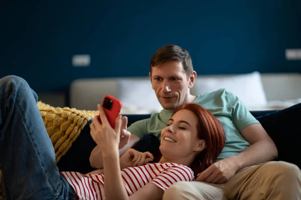Couple in love resting on comfy couch having fun using smart phone apps, enjoy distant video call, wife showing interesting website to husband, choosing services good, married couple shopping online