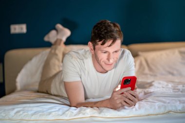 Man reading information in smartphone lying in bed at home in modern bedroom in evening. Interested concentrated male searching info, surfing online, watching content, scrolling social media in phone. clipart