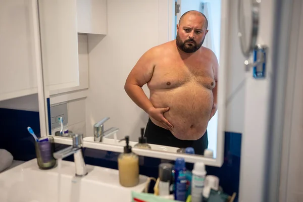 stock image Dissatisfied with weight man stands in bathroom looking at big belly from overeating in mirror. Frustrated fat man holding huge abdomen thinking about problem of excess weight, new diet for slimming.