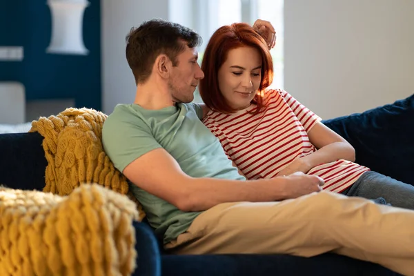 Loving woman looking at caring man with trust in close relations. Wife and husband sitting on cozy sofa gentle hold hands, have sincere feelings, look at eyes each other. Romantic weekend at home.