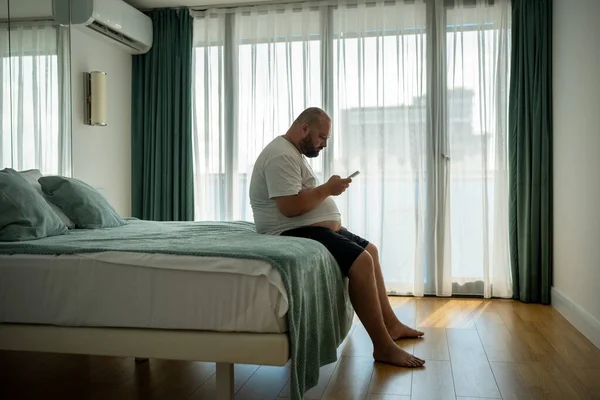 Chlápek Nadváhou Visící Smartphonu Letovisku Sedící Posteli Hotelovém Pokoji Dovolené — Stock fotografie