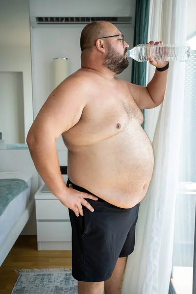 stock image Excess weight man drinking water in air-conditioned hotel room looking at window in hot weather. Overweight guy with naked torso suffers from heat. Heat stroke, overheating, unhealthy body shape. 