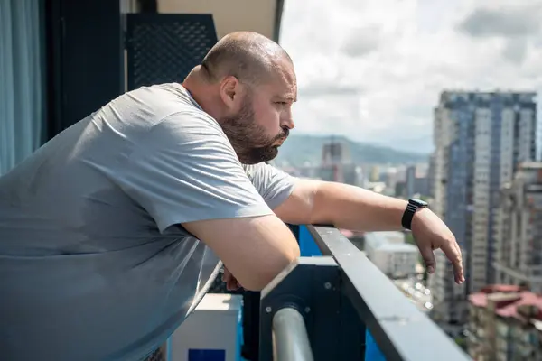 Overweight man feeling bad from heat in summer standing on balcony looking at street in summer. High sweating, hyperhidrosis, obesity, heaviness from hot weather high humidity, excess body weight.