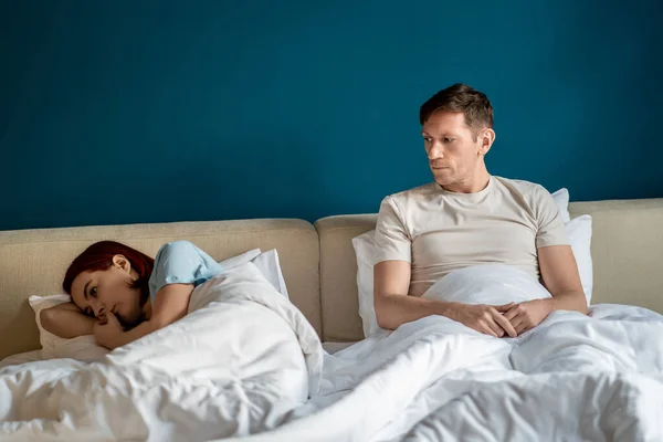 Husband tries to talk to offended wife in bed at home sitting near covering blanket. Woman ignoring man turning away from him. Frustrated spouses having misunderstanding, ending of marriage, divorce.
