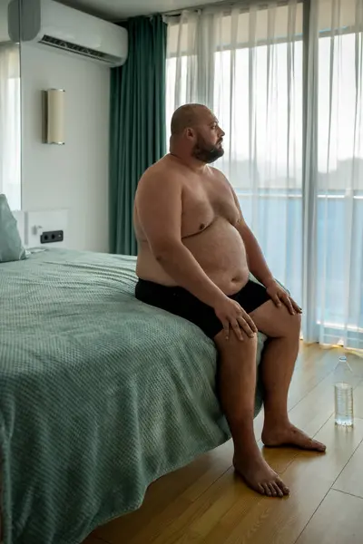 Overweight man in shorts sitting in bed in hotel room looking at window on vacation. Obesity male plump chubby guy suffer from hot, stuffiness resting on resort. Health problem of excess weight.