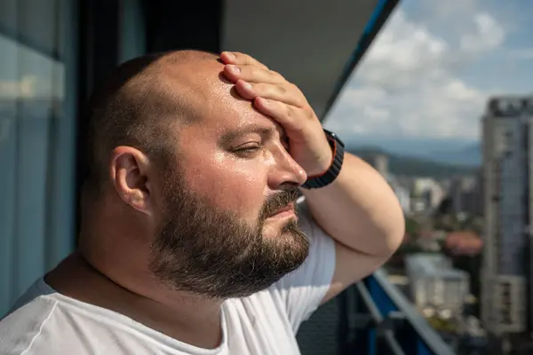 stock image Portrait overweight man has headache touching forehead in heat summer weather. Bearded guy with excess weight looking from balcony on street in city. Extra high temperature, health problems concept.