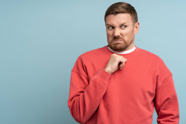 Confused man feeling disgust with serious face incredulously looking aside isolated on blue background. Clenching fist gesture on chest. Guy has scared expression, demonstrates aversion makes grimace.