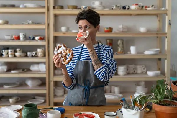Young creative tattooed female potter in dirty apron drinking tea from handcrafted clay mug and eating tasty pretzel at workshop. Portrait of modern ceramics master at workplace