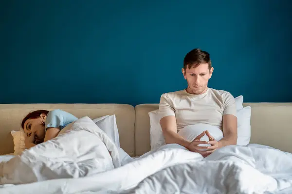 Thoughtful sad man woman in bed feeling despair after quarrel at home. Husband sits looks at hands, wife turned away lying near. Misunderstanding, problem relationships, marital discord, ignoring.