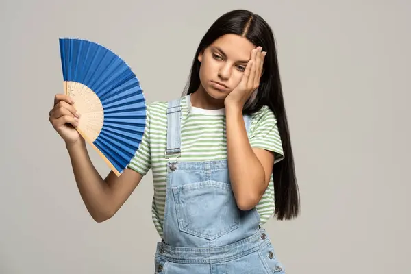 stock image Too hot. Sweaty tired teenage girl touching forehead using paper fan suffer from heat, feels sluggish. Displeased teen girl cooling in hot summer weather, isolated on studio gray wall. Overheating