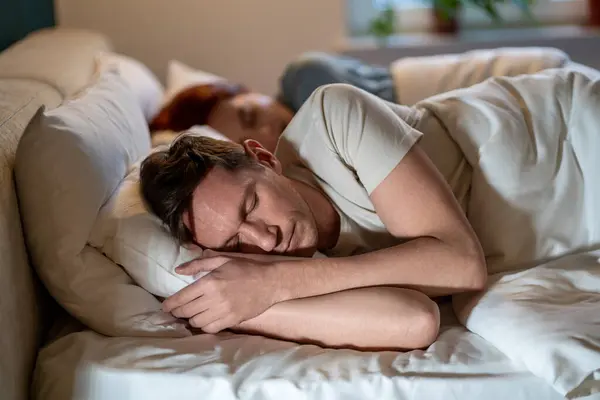 Man and woman sleep soundly well. Guy and girl dozing on same bed on soft regular comfortable pillows under warm blankets in glow of lights. Importance of long, healthy sleep. Joint sleep of spouses