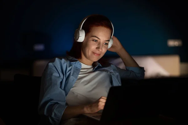 Woman with laptop watches video with headphones. Calm girl happily smiles quite broadly using computer, watches funny movie, useful webinar, spends time online on Internet late at night in dark room