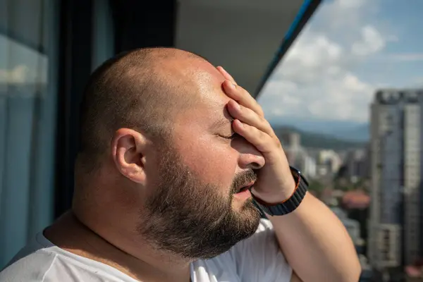 Portrait overweight man has headache touching forehead in heat summer weather. Bearded guy with excess weight closed eyes stand on balcony. Extra high temperature, health problems concept.