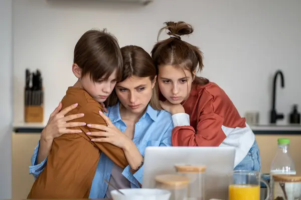 Teenagers children support mother in kitchen looking at laptop screen with bad news from work. Boy girl hug mom with sad faces. Single mother raising son daughter working online, family care concept.