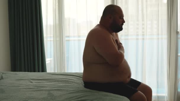 中年男人为了应付夏天的烦闷 经常做呼吸练习 肥胖肥胖的胖胖的年轻男性坐在床上 呼吸急促 闷热难忍 抱着胸部 — 图库视频影像