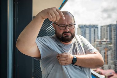 Man with wet armpits and disgusted expression on face. Fat obese young guy with glasses suffering from severe sweating, overweight. Humid climate and high air temperature in city in hot summer. clipart