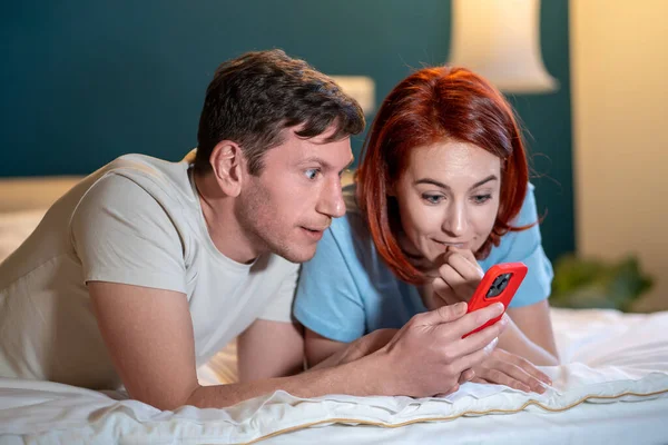 Happy marriage couple lyimg on bed talking reading good news on phone screen. Excited wife and husband receive important message on cell together. Overjoyed man and woman surprised win online lottery