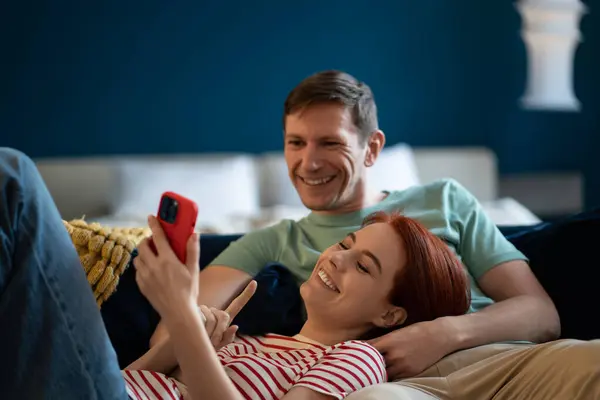 Happy rested man and pleased woman lying relaxed on sofa look screen cellphone. Positive family online shopping Smiling middle aged married couple chatting with friends via video call, social network