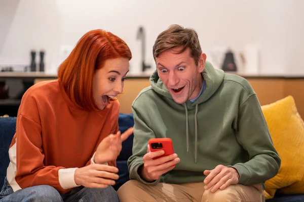 Amazed shocked happy man woman rejoicing looking at smartphone screen at home. Winning the lottery, good news, financial profit, success in family business, sales in online shop, store, entrepreneurs.
