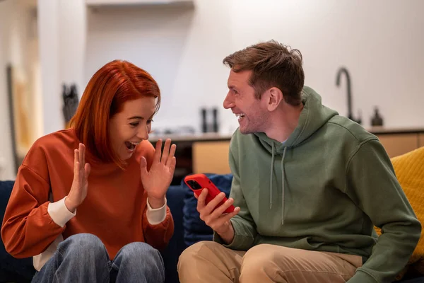 Happy overjoyed couple man woman reading good news in smartphone sitting on couch at home. Wife husband smiling rejoicing together, gesturing hands, having amazed wondered faces.