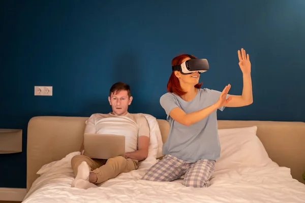 Woman in 3D goggles immersive in virtual world while man with laptop work in bed different spend time at home. Wife sits in augmented reality glasses play video games. Technologies for entertainment