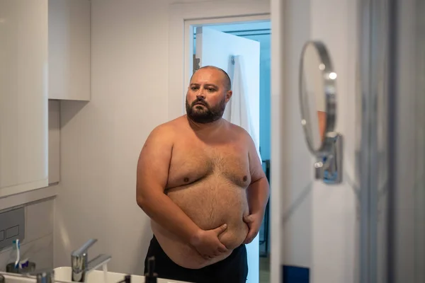Upset man has extra weight looking at mirror touching big abdomen in bathroom at home. Bearded guy dissatisfied body shape. Obesity, overweight, healthy problems, weight control, bad unhealthy habits.