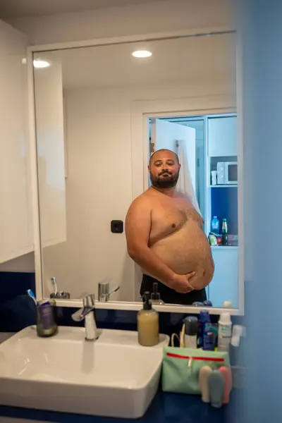 stock image Overweight smiling man with naked torso suffering from extra weight looking at mirror touching abdomen in bathroom at home. Bearded middle aged male inspecting body to change unhealthy life habits. 