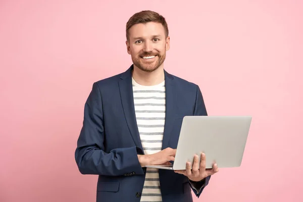 stock image Positive man trader, crypto investor, entrepreneur, financier holds laptop on pink background. Bearded well groomed smiling guy wearing jacket, jeans looking at camera on advertisement banner poster.