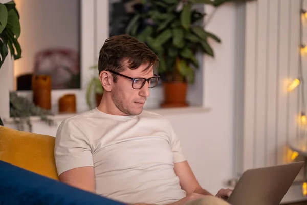 Man sits on couch with laptop on lap. Middle aged guy with glasses in cozy apartment in evening works overtime at computer, completes online Internet IT project, remote studying work from home