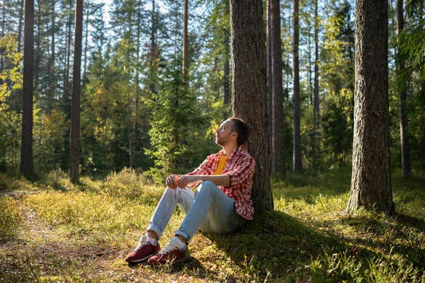 stock image Man camper tourist resting sitting near tree in forest with closed eyes in warm sunny day. Guy enjoying in unity with nature in park. Male relaxing dreaming meditating, tourism travel hiking concept.