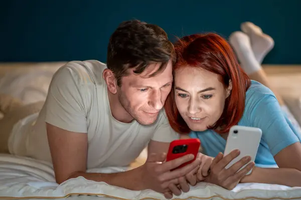 Married couple uses phones together. Middle aged man woman lie on bed on stomachs watch interesting fascinating videos, check news on Internet social networks. Joint leisure time of romantic couple