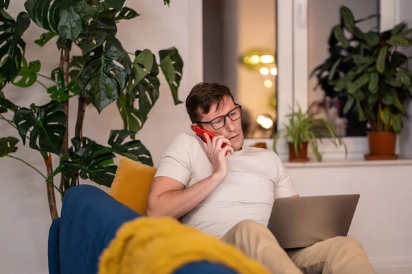 Man in eyeglasses talks phone works on laptop sits on couch at home in evening. Discuss project, working issues, tasks, problems with colleagues. Home office, distant, remote, online job work concept.