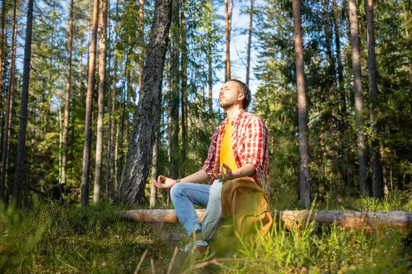 Relaxed man sitting in lotus position meditating in sunlight while resting on timber after long hiking. Active pastime, healthy lifestyle, physical activity, energy balance recreation in nature