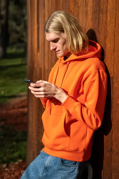 Handsome relaxed confident guy with long hair leaning on wooden wall. Brooding caucasian man in orange sport hoodie looks into mobile telephone, writes message, plays online games, reads news, article
