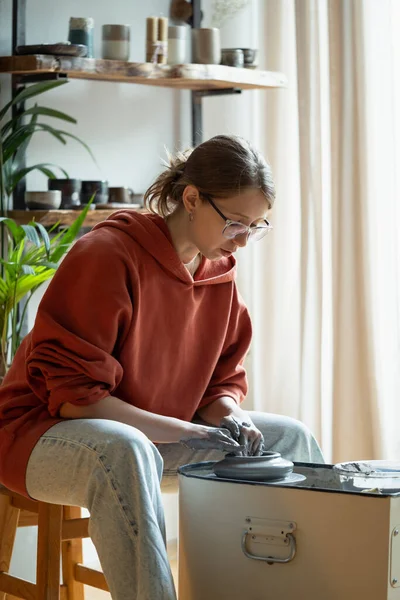 Stock image Relaxed woman woman making natural clay utensils as hobby to relieve stress after work week. Carefree girl learning pottery sits in ceramic workshop at table with spinning stand and pedal