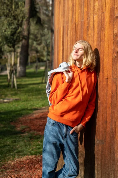 Relaxed blond long hair guy scandinavian androgynous appearance standing near wooden fence in park with closed eyes. LGBT, gay, transgender man walks outdoor enjoying nature. Rest, relax, leisure.